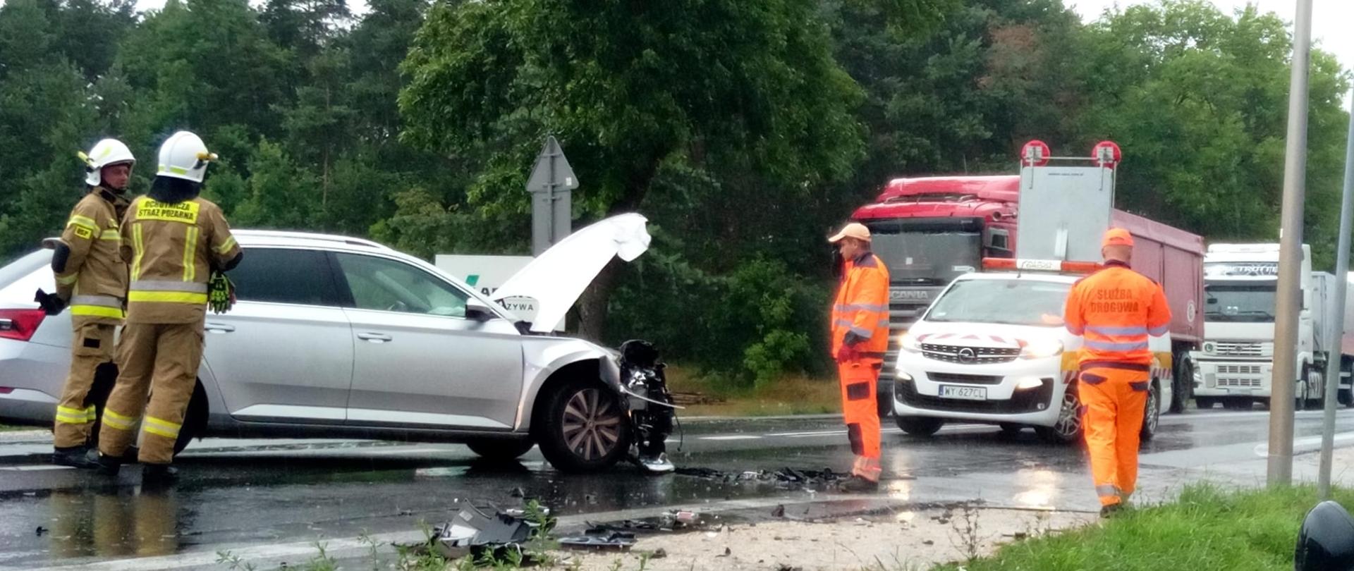 Zdjęcie przedstawia drogę krajową. Po prawej stronie zniszczony samochód osobowy przy którym stoi dwóch strażaków. W centralnym punkcie, w pomarańczowych uniformach przedstawiciele zarządcy drogi, przy nich ich samochód. Dalej widać przypadkowe pojazdy stojące na pasie drogowym zamkniętej ulicy. 