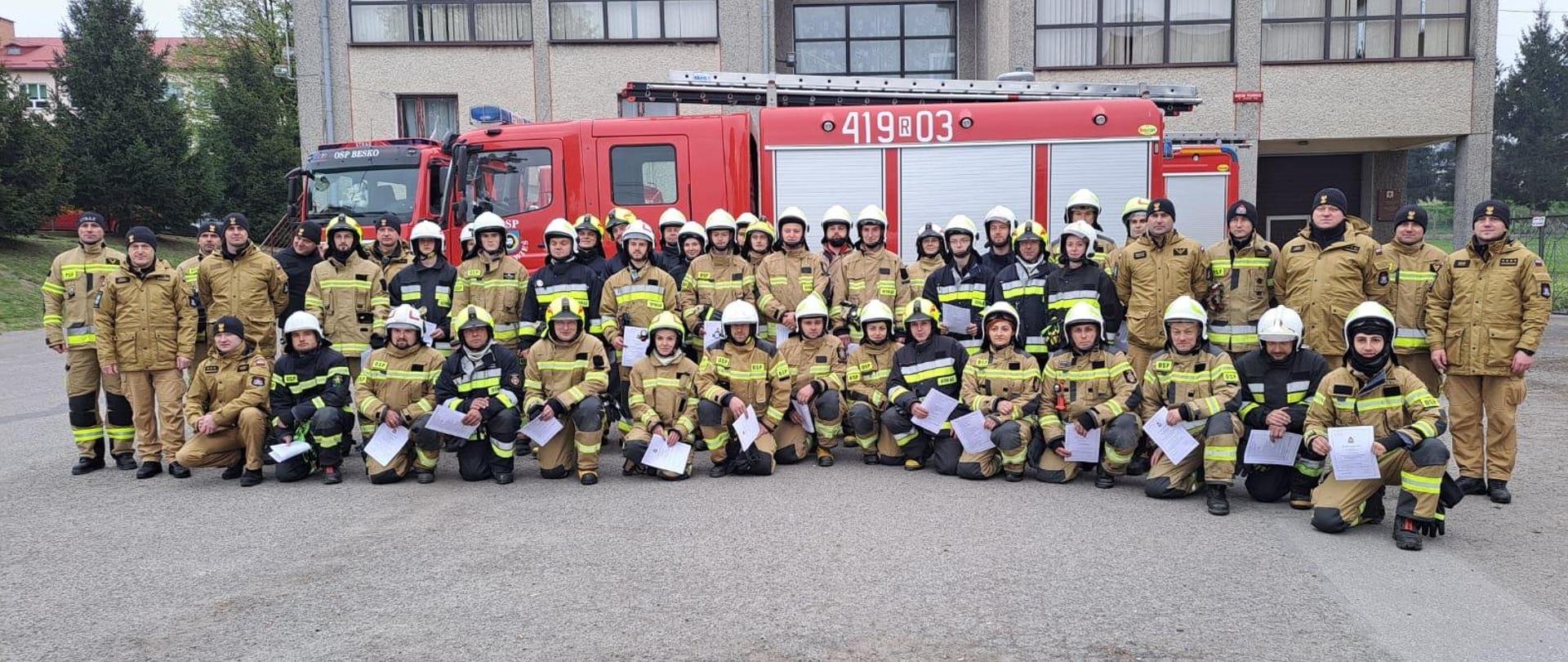 Kolorowa fotografia wykonana na zewnątrz w pochmurny dzień. Przedstawia strażaków OSP w ubraniach specjalnych i komisję sędziowską PSP w ubraniach służbowych na podsumowaniu szkolenia podstawowego strażaka OSP.