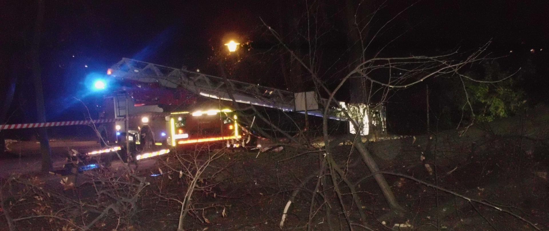 Zdjęcie przedstawia połamane drzewo oraz porozrzucane gałęzie - w tle widać strażacki podnośnik na sygnałach oraz otaśmowany teren działań strażaków.