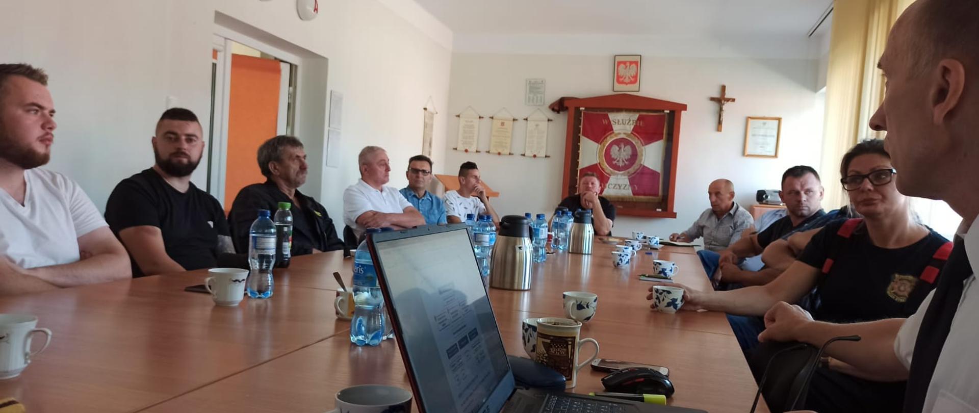 Zdjęcie przedstawia grupę ludzi siedzących w świetlicy przy stołach, którzy słuchają prowadzącego spotkanie ubranego w białą koszulę oraz czarny krawat. W tle widzimy na ścianie godło Polski, sztandar KP PSP Świdwin oraz krzyż.