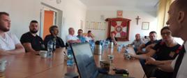 Zdjęcie przedstawia grupę ludzi siedzących w świetlicy przy stołach, którzy słuchają prowadzącego spotkanie ubranego w białą koszulę oraz czarny krawat. W tle widzimy na ścianie godło Polski, sztandar KP PSP Świdwin oraz krzyż.