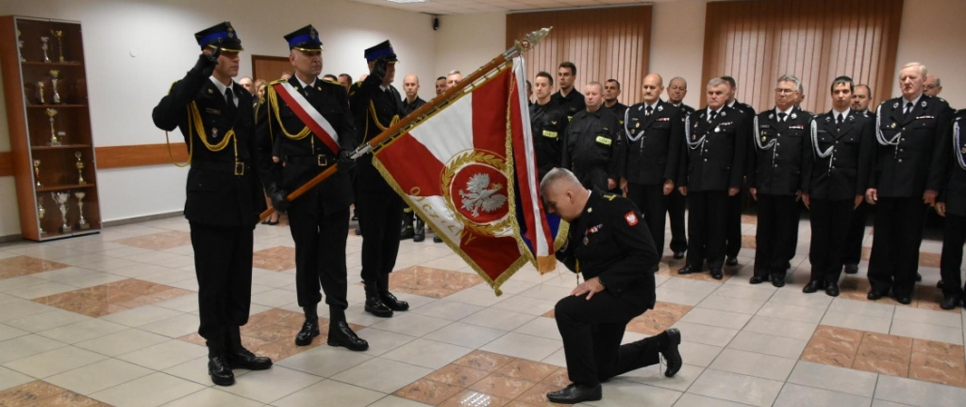 Komendant powiatowy uklęka i całuje sztandar Komendy Powiatowej PSP w Skarżysku-Kamiennej. Przed nim stoi poczet sztandarowy. W tle widać druhów OSP i funkcjonariuszy PSP.