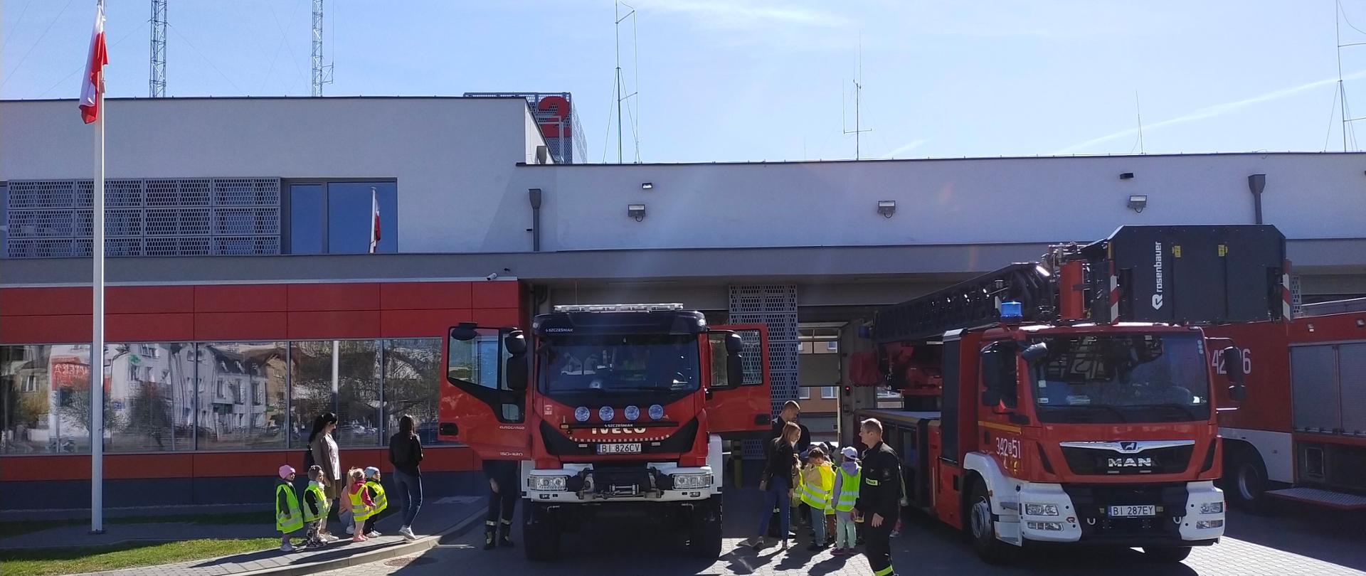Na tle budynku Komendy Miejskiej PSP widać pojazdy pożarnicze a przy nich dzieci z opiekunami i strażakami