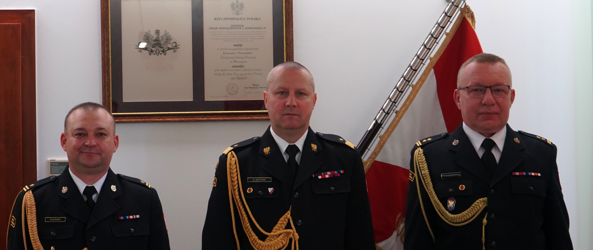 trzech strażaków w mundurach ze sznurem , w tle sztandar