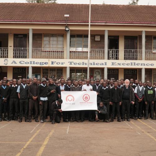 Grupa ok. 100 osób pozuje do zdjęcia przed szkołą trzymając przed sobą plakat z logo polskiej pomocy i SWM