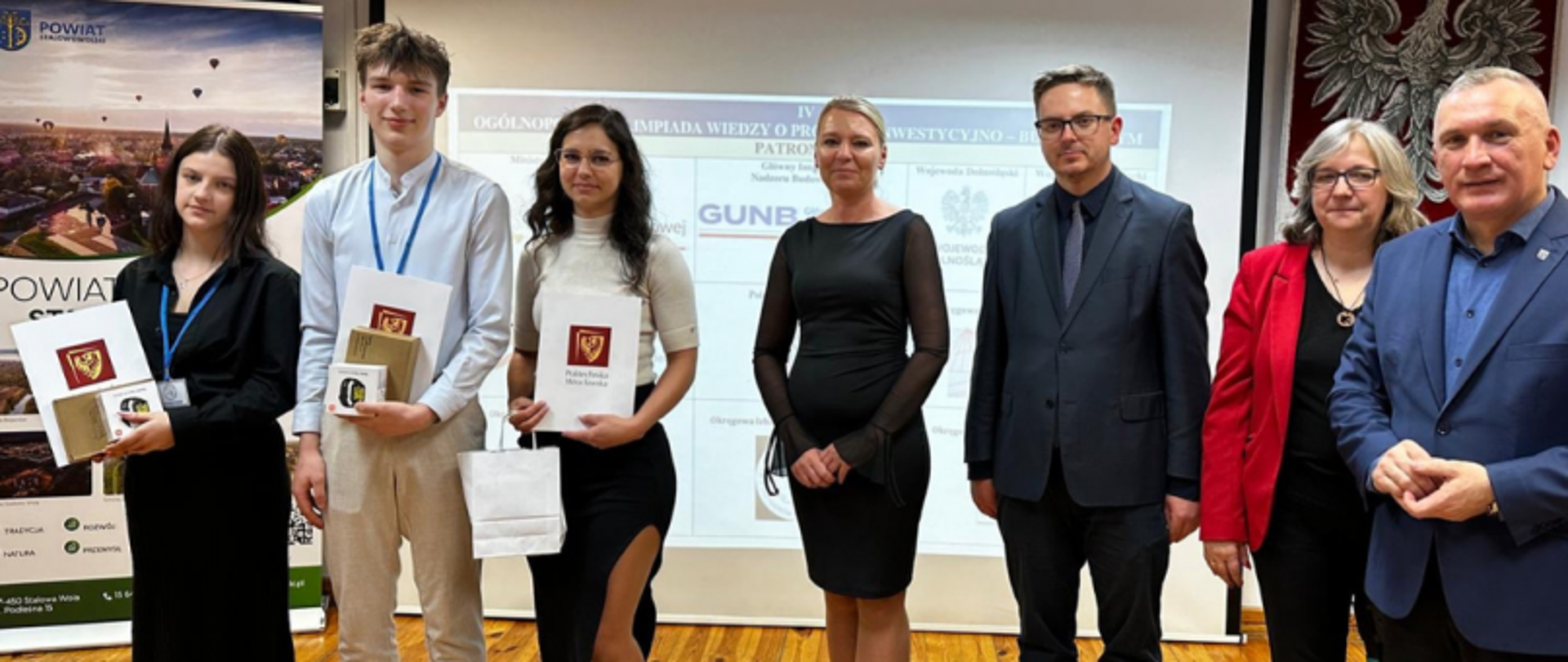 Zwycięzcy i organizatorzy podczas finału IV Ogólnopolskiej Olimpiady Wiedzy o Procesie Inwestycyjno-Budowlanym; młodzież trzyma dyplomy i nagrody, w tle widoczne są banery i ekran z logotypami patronów wydarzenia, w tym Głównego Urzędu Nadzoru Budowlanego (GINB).