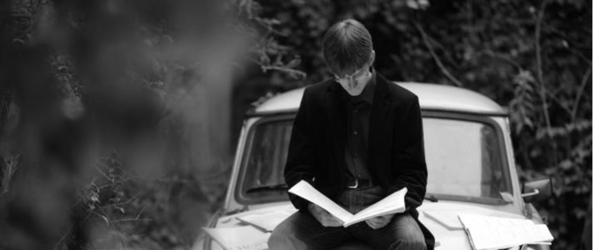 w centralnej części czarnobiała fotografia przedstawiająca mężczyznę z książką siedzącego na masce samochodu, powyżej i poniżej informacje dotyczące spotkania z Marcinem Koziakiem 