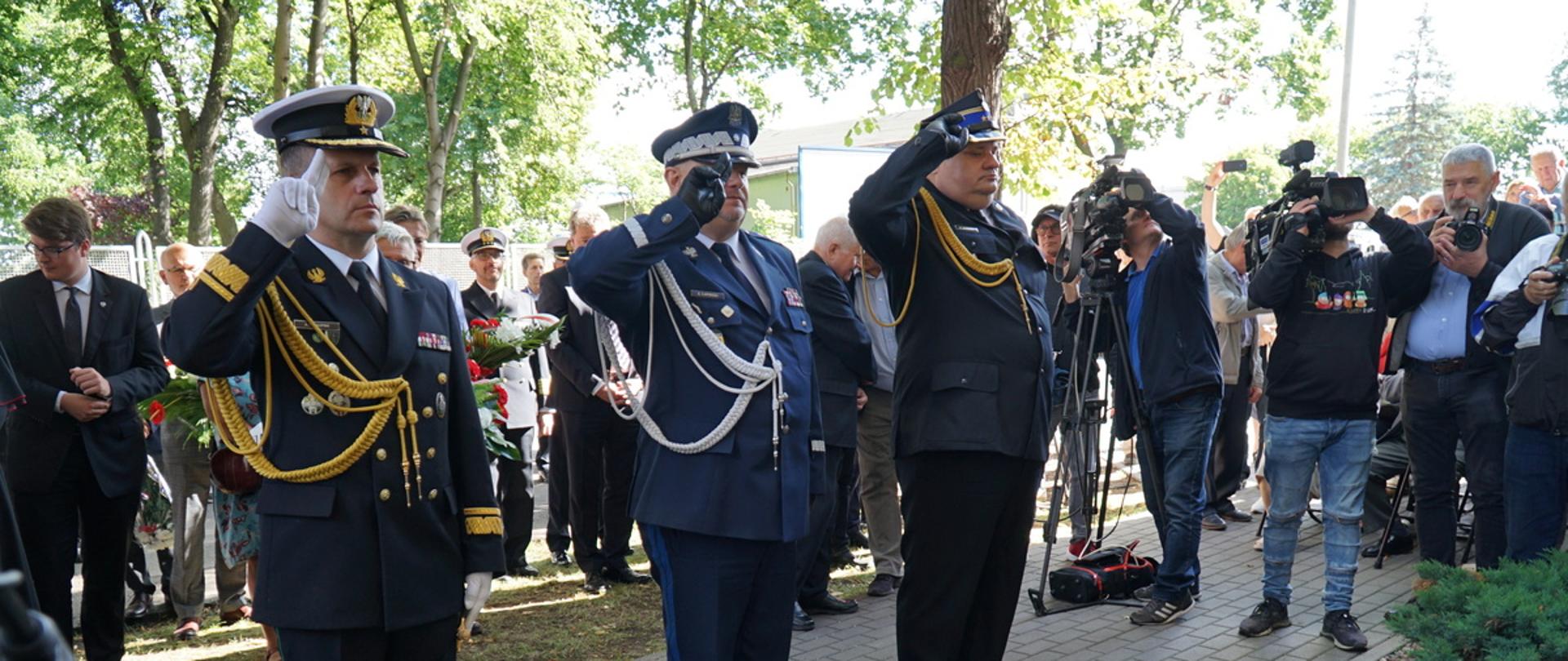 Funkcjonariusze Straży Granicznej Policji oraz Państwowej Straży Pożarnej stoją oddając honor za nimi stoją zaproszeni goście.