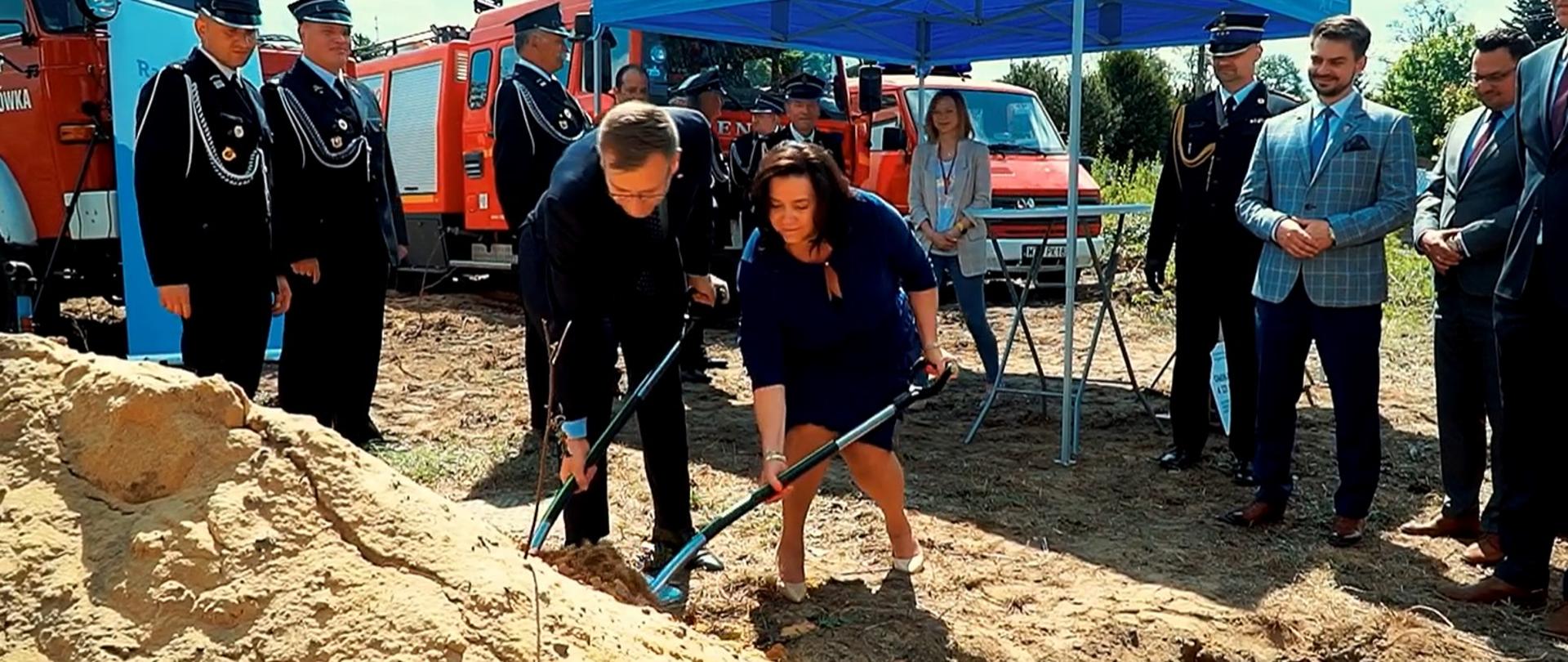 Wójt gminy Radziejowice Urszula Ciężka i sekretarz stanu w Ministerstwie Aktywów Państwowych Maciej Małecki wbijają łopaty w hałdę piasku. Wokół nich stoją ludzie. W tle stoją strażackie wozy bojowe.