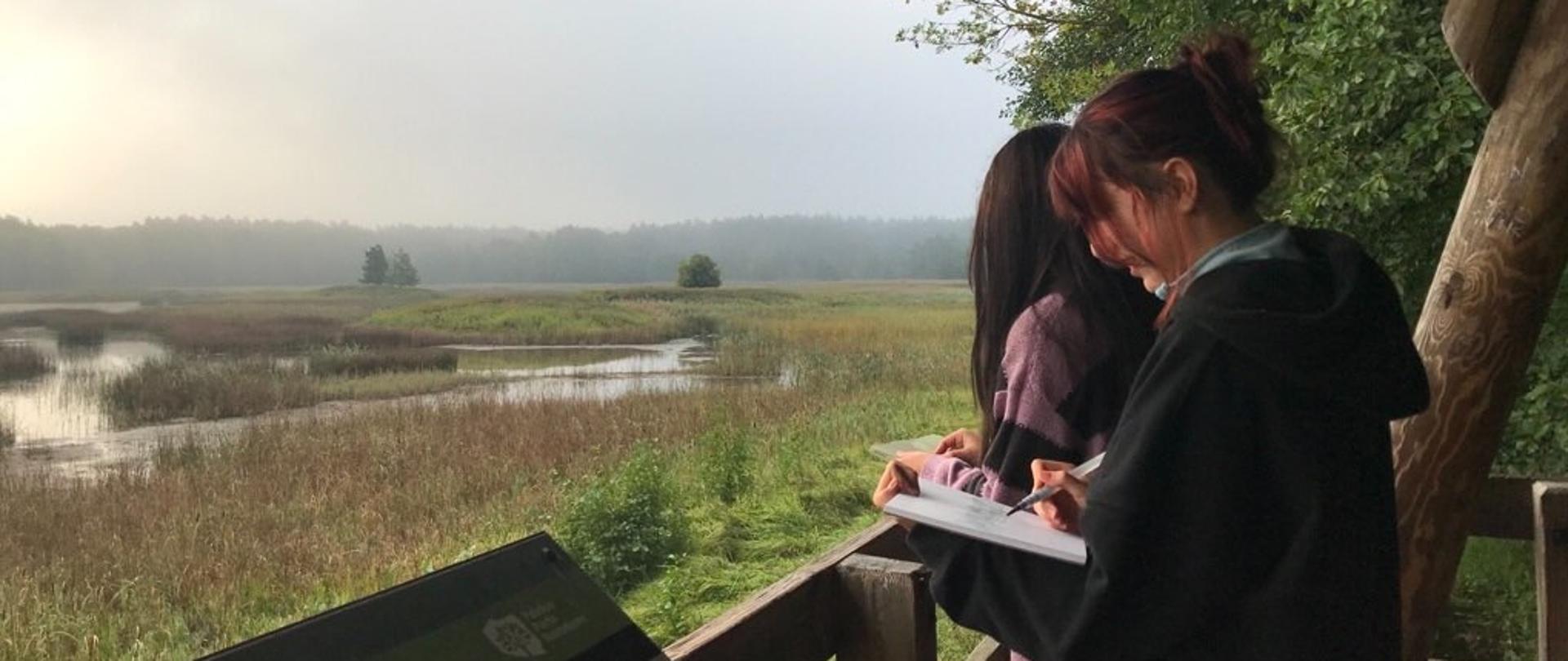 zdjęcie ,taras widokowy dwie dziewczynki szkicują pejzaż , rozlewisko rzeki w oddali las