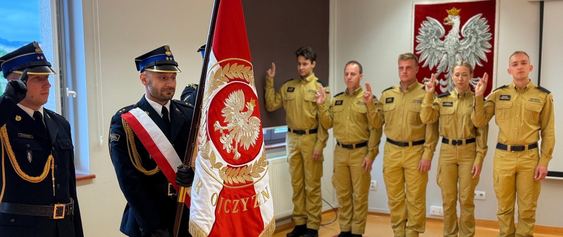 W sali z jasną podłogą, przed ścianą z godłem Polski, stoi grupa strażaków. Na pierwszym planie, centralnie, strażak w ciemnogranatowym mundurze z biało-czerwoną szarfą trzyma rozwinięty sztandar z godłem i napisem "OJCZYZNA". Po jego lewej stronie inny strażak w ciemnym mundurze salutuje. W tle, w rzędzie, stoi pięciu młodych strażaków w piaskowych mundurach, z których czterech trzyma uniesione dwa palce prawej ręki w geście przysięgi.