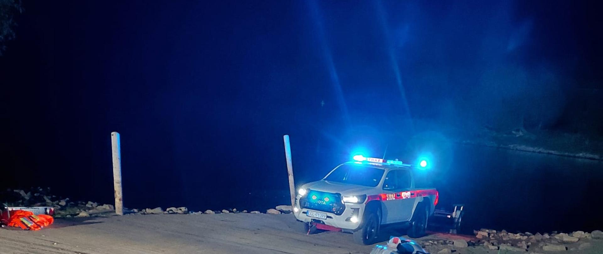 Nocne działania ratownicze nad zbiornikiem wodnym. Na pierwszym planie znajduje się nosze transportowe przygotowane do udzielenia pomocy poszkodowanemu. W tle widoczny jest samochód ratowniczy Państwowej Straży Pożarnej z włączonymi sygnałami świetlnymi, ustawiony przy pochylni prowadzącej do wody. Miejsce zdarzenia jest oświetlone przez pojazdy ratownicze.