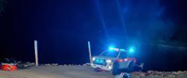 Nocne działania ratownicze nad zbiornikiem wodnym. Na pierwszym planie znajduje się nosze transportowe przygotowane do udzielenia pomocy poszkodowanemu. W tle widoczny jest samochód ratowniczy Państwowej Straży Pożarnej z włączonymi sygnałami świetlnymi, ustawiony przy pochylni prowadzącej do wody. Miejsce zdarzenia jest oświetlone przez pojazdy ratownicze.