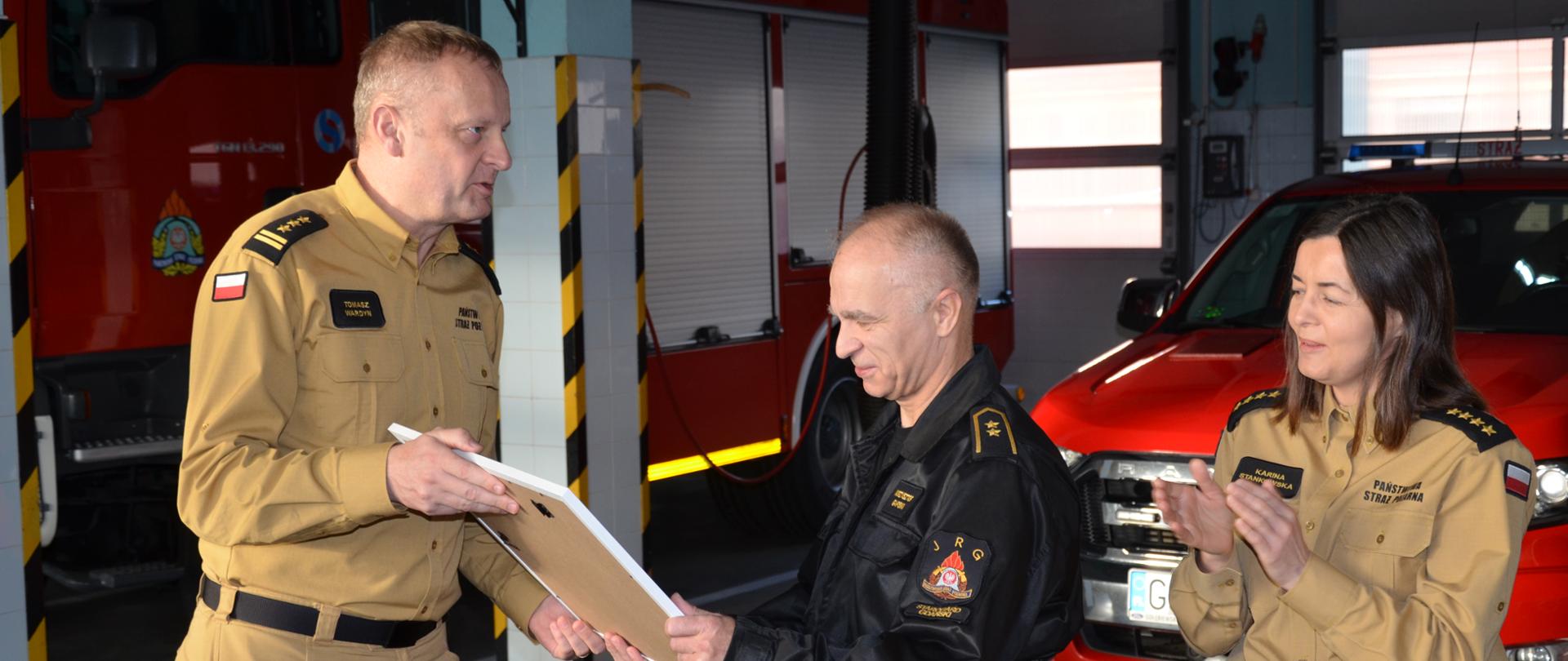 Na zdjęciu komendant powiatowy wręcza pamiątkowe zdjęcie funkcjonariuszowi, w tle samochody pożarnicze