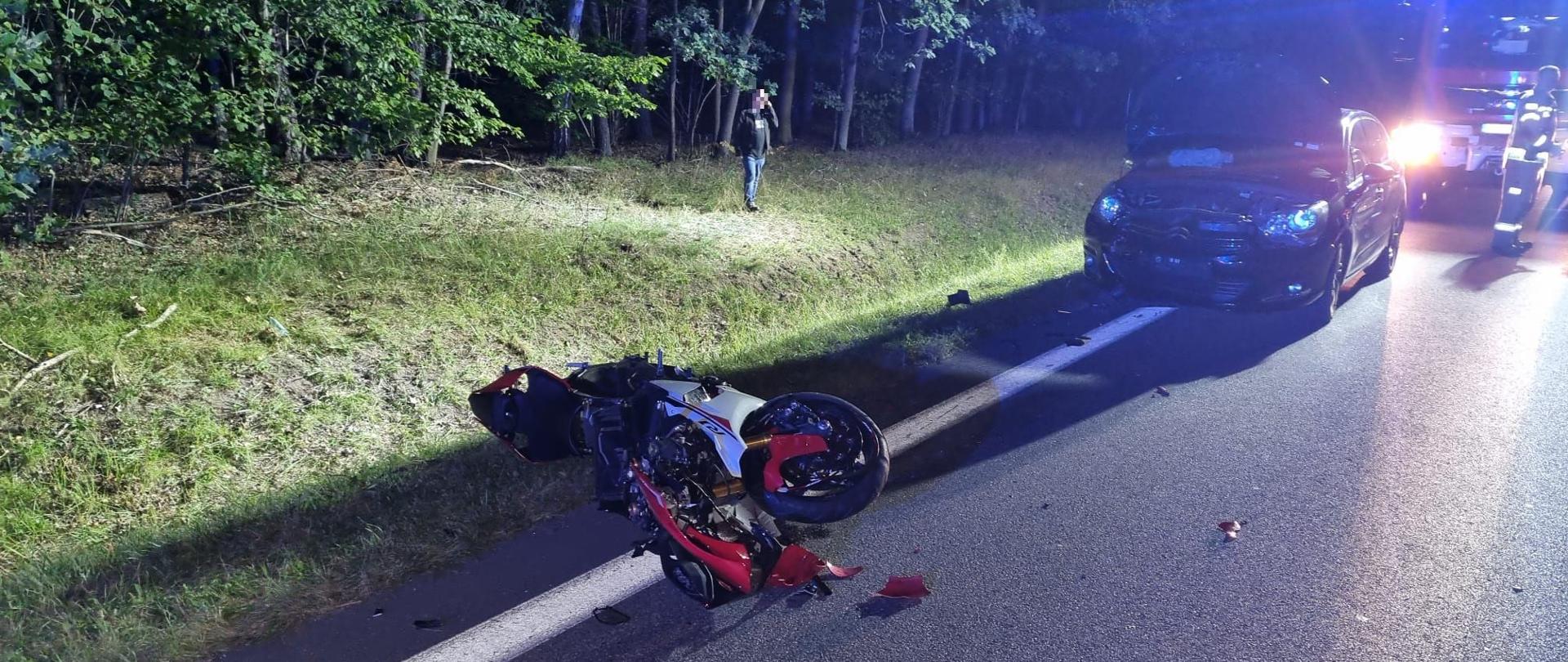Na pierwszym planie w światłach samochody strażackiego widać roztrzaskany motocykl, na drugim planie samochód osobowy z uszkodzonym przodem.