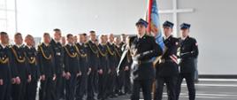 Trzech strażaków ze sztandarem idzie przed strażakami w umundurowaniu wyjściowym ze sznurem stojącymi obok siebie w rzędzie.