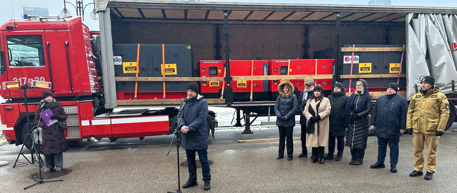 Wywiadu udziela Prezydent Miasta Stołecznego Warszawy Rafał Trzaskowski. Po lewej stoi Rzecznik Prasowa,a po prawej inni uczestnicy konferencji. Tło zdjęcia stanowi otwarta naczepa wypełniona skrzyniami z agregatami. 