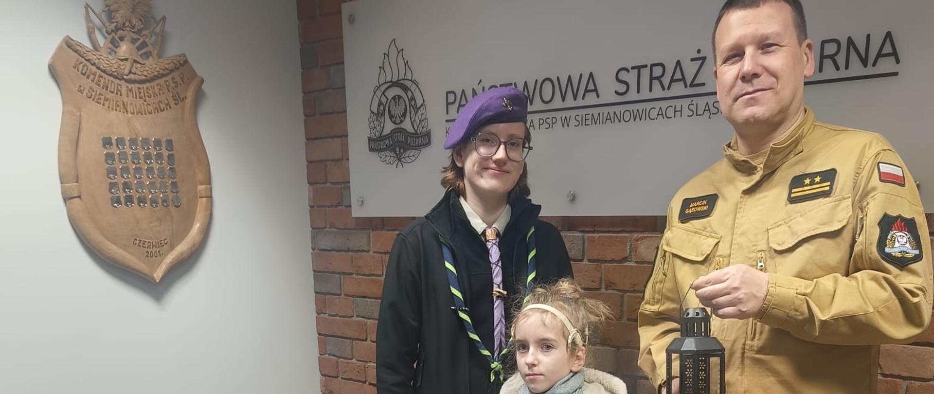 Dwójka harcerzy — starszy i młodsze dziecko — przekazuje Betlejemskie Światło Pokoju Komendantowi Miejskiemu PSP w Siemianowicach Śląskich. Komendant trzyma lampion z płomieniem. W tle ceglana ściana, tablica z logo PSP oraz świąteczne dekoracje ustawione na stole.