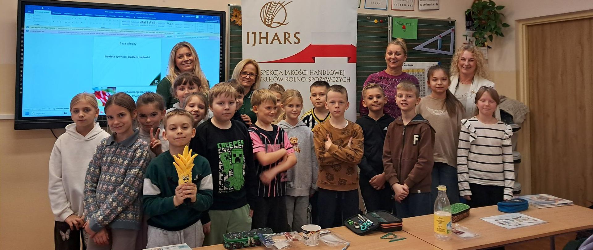 Grupa uśmiechniętych dzieci stoi w klasie razem z inspektorkami IJHARS i nauczycielkami po zakończonych zajęciach. W tle widać szkolne ławki, tablicę i kolorowe dekoracje. Dzieci trzymają maskotkę IJHARS.