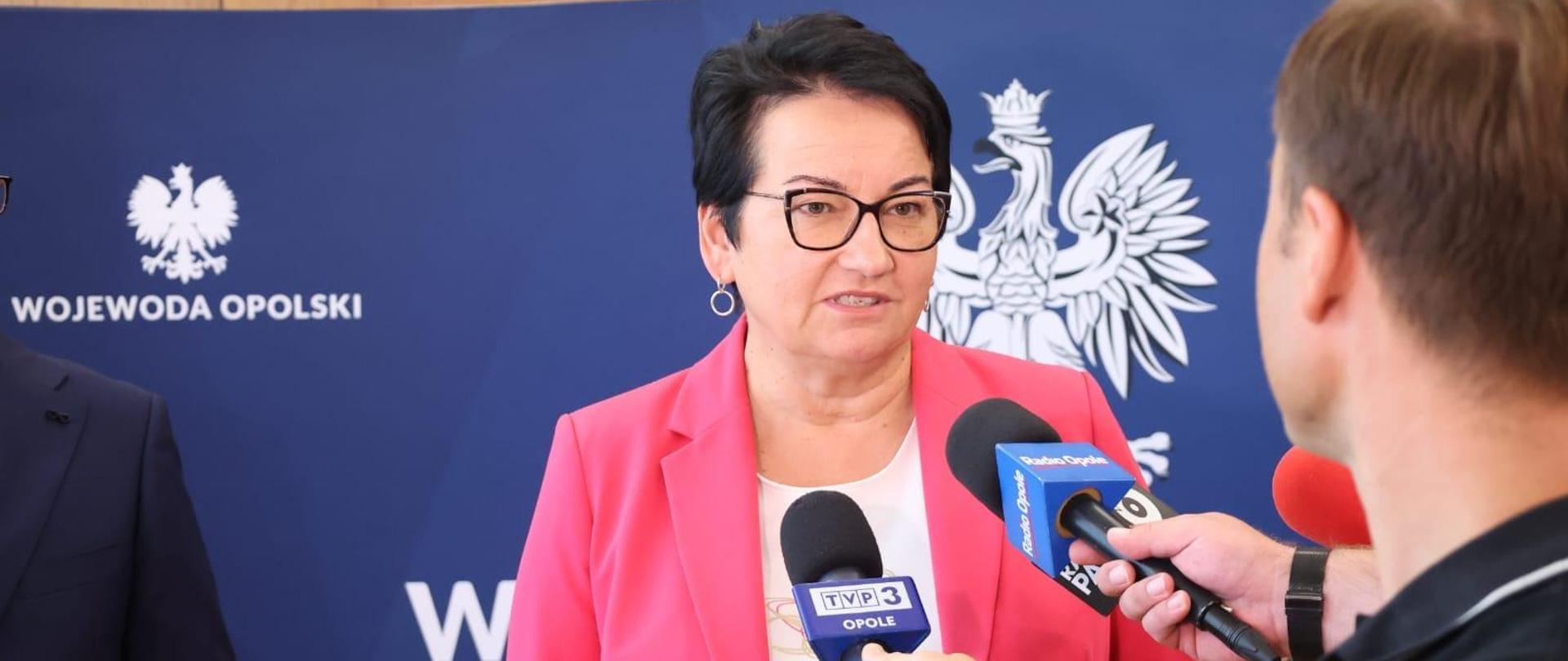 Wojewoda mówi do mikrofonów trzymanych przez dziennikarzy. Widzimy ją od pasa w górę. Patrzy na jednego z dziennikarzy. Widzimy tylko tył jego głowy. 