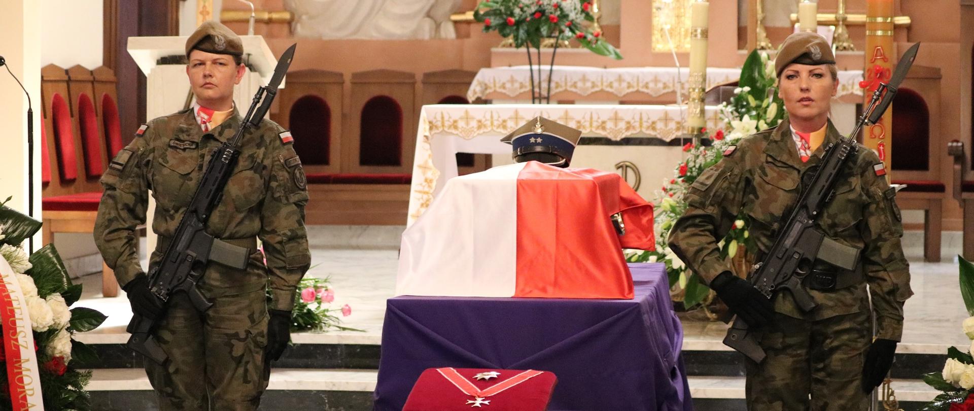 Dwie kobiety - żołnierze stoją obok urny udekorowanej biało-czerwoną flagą.