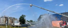Na fotografii: ramię dźwigu wraz ze strażakiem nań stojącym, strzelający wodom w ramach symulowanej akcji gaszenia wycieku łatwopalnego surowca ze zbiornika