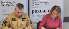 Strażak oraz kobieta siedzą przy stole podpisując dokumenty. Za nimi znajdują się banery.