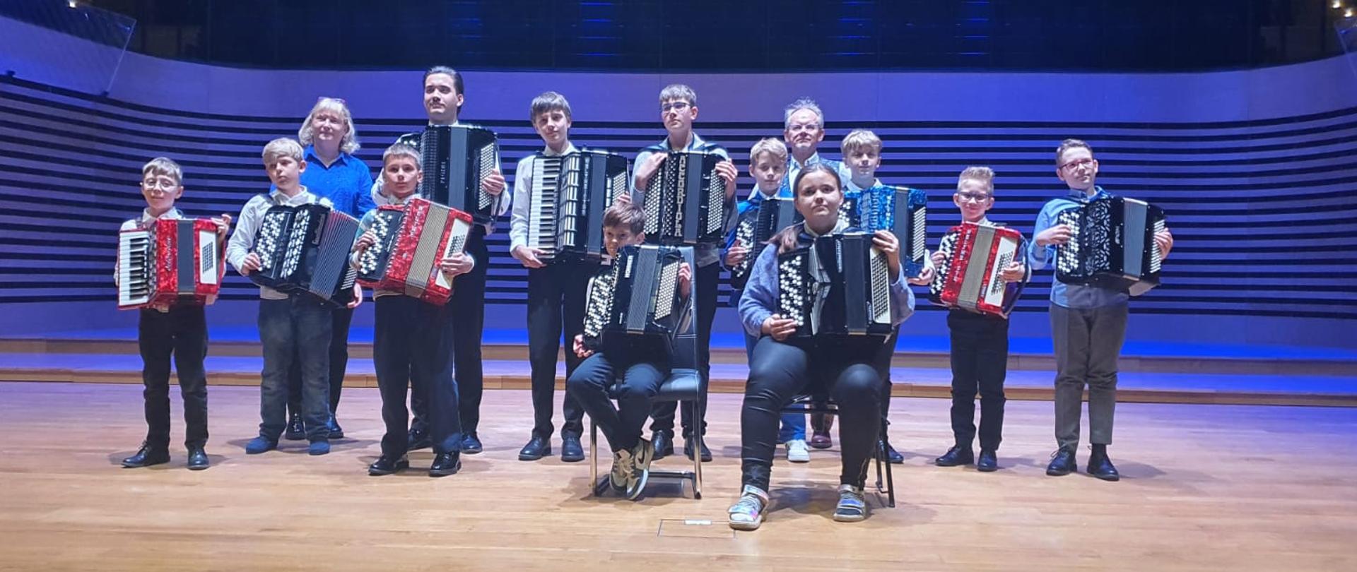 Kobieta i mężczyzna oraz wszyscy wykonawcy stoją na scenie z akordeonami.