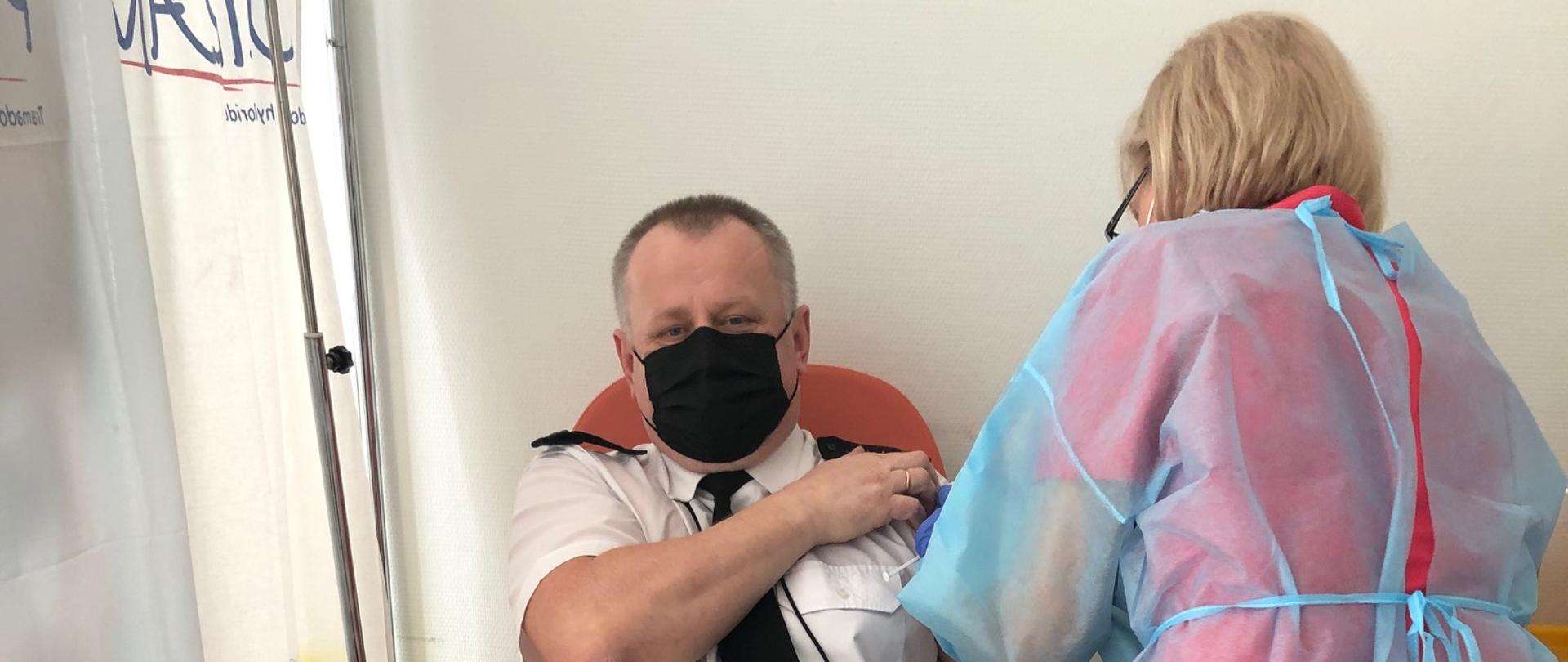 Strażak siedzący na fotelu jest szczepimy przez pielęgniarkę w lewą rękę.