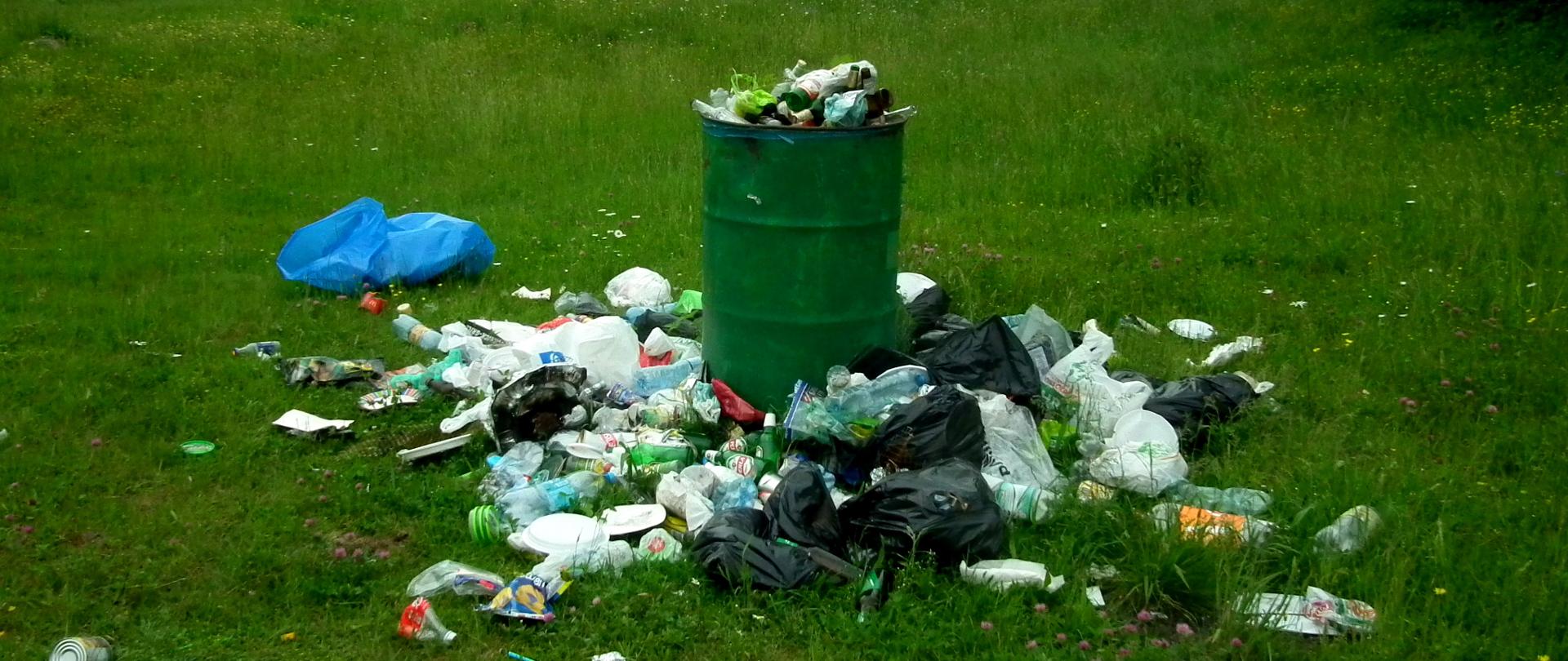 Na łące stoi beczka pełna śmieci. Śmieci leżą też wokół. 