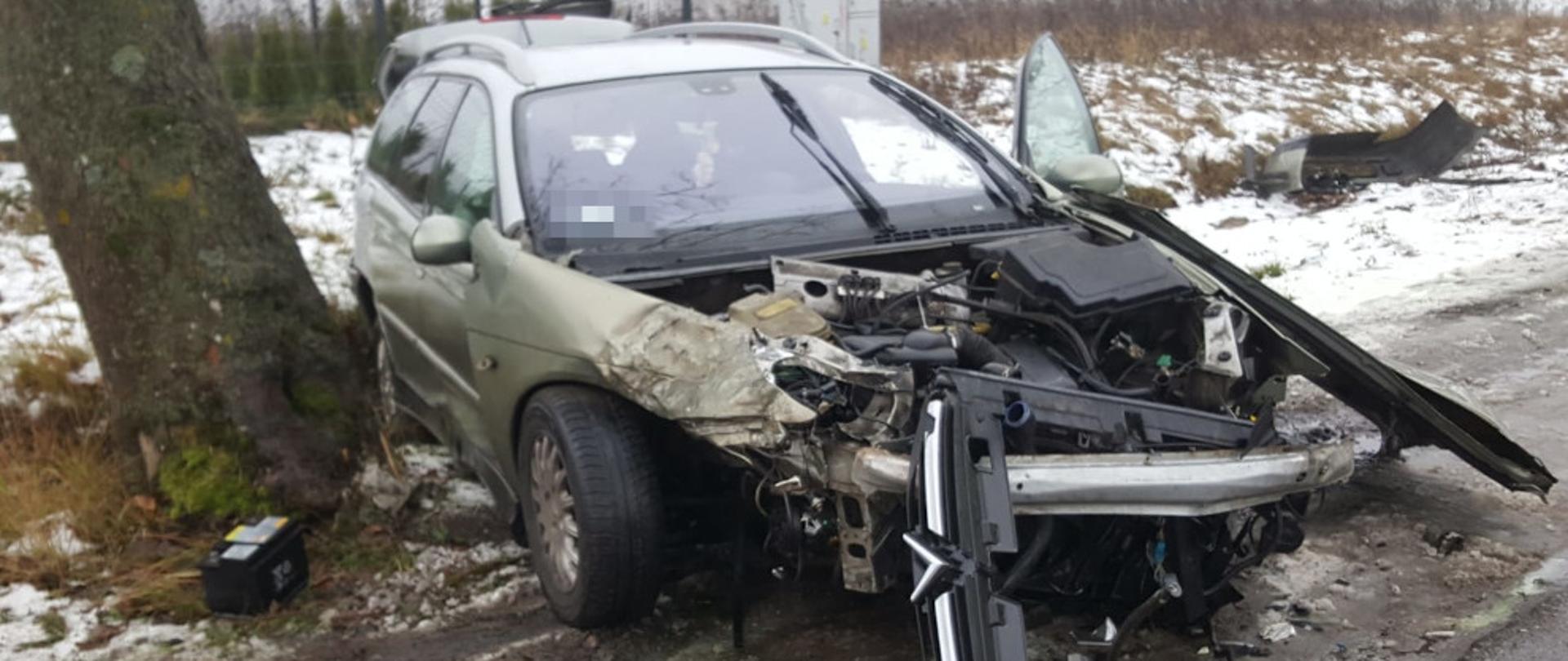 Rozbity samochód marki citroen, uszkodzony przód pojazdu. Stoi na poboczu (prostopadle do drogi). Obok pojazdu leży akumulator.