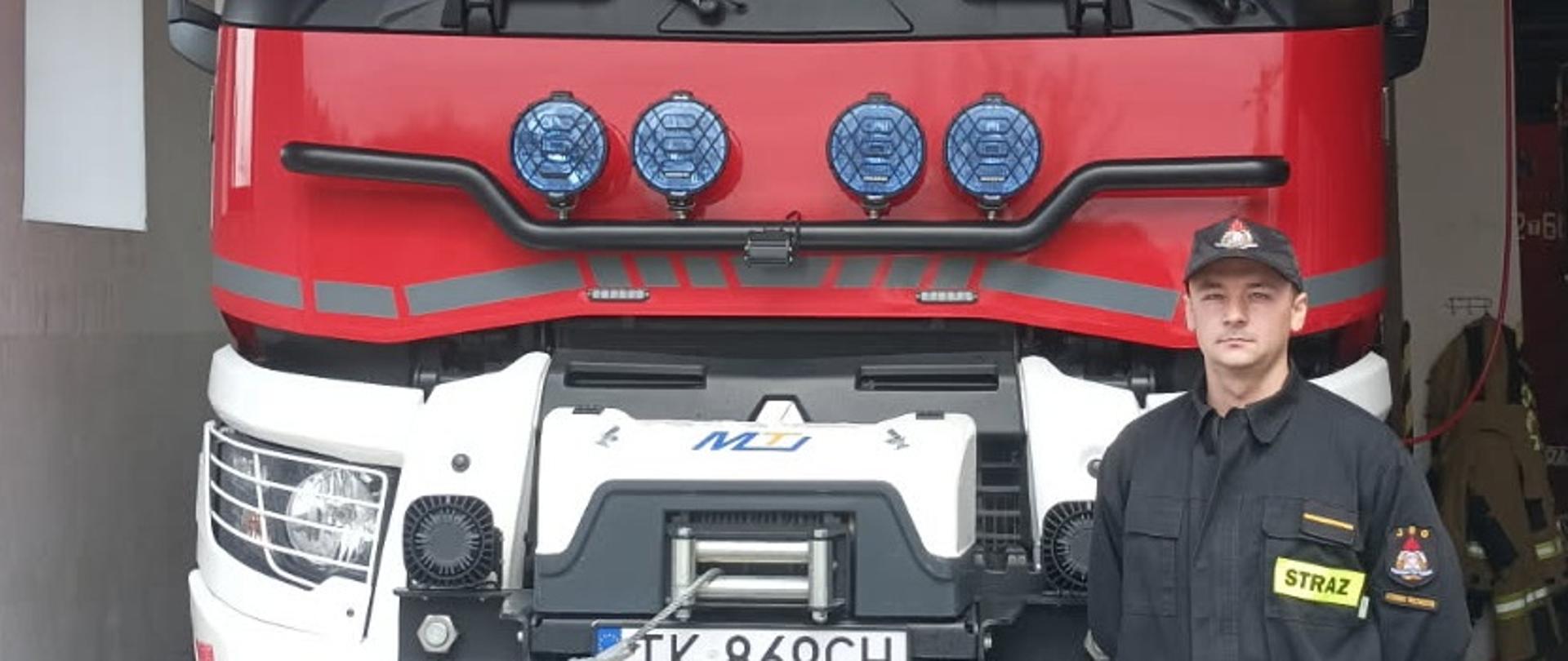 Na zdjęciu widać strażaka, który stoi przed samochodem strażackim zaparkowanym w remizie.