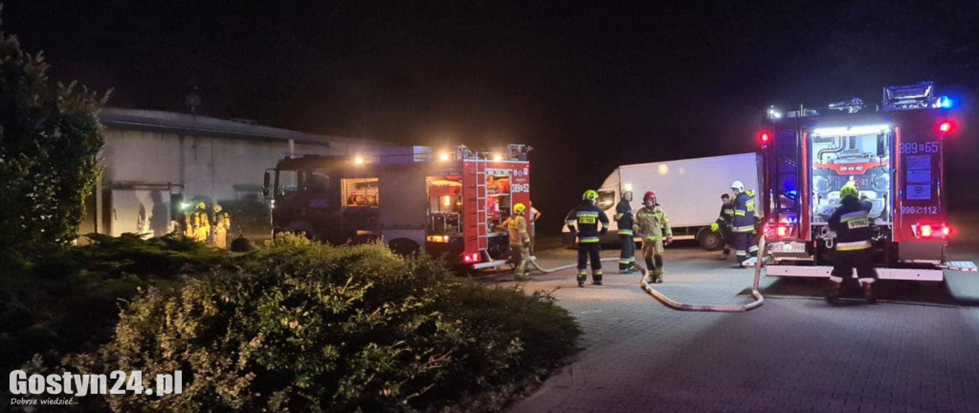 Strażacy prowadzą działania na placu przy zakładzie. Na zdjęciu znajdują się także dwa samochody pożarnicze.