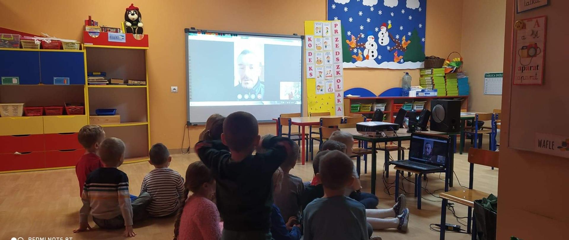 Na zdjęciu widoczna jest sala lekcyjna w przedszkolu. Na pierwszym planie, w centralnej części fotografii widoczna jest grupa dzieci – dziewczynek oraz chłopców, którzy siedzą na podłodze. Wszyscy zwróceni są twarzami w kierunku ekranu, na którym wyświetlany jest strażak, podczas prowadzonej na żywo transmisji. Ekran zawieszony jest na jednej ze ścian sali, w jej centralnej części. Po lewej stronie ekranu, przy ścianie widoczne są kolorowe meble na pomoce dydaktyczne. Po prawej stronie ekranu, przy ścianie widoczne są kolorowe regały, na których zgromadzone są materiały edukacyjne. Powyżej widoczna jest dekoracja zimowa w formie gazetki oraz kodeks przedszkolaka. Pomiędzy dziećmi a regałami znajdują się stoliki oraz krzesełka dziecięce. Na jednym ze stolików ustawiony jest rzutnik multimedialny, który wyświetla obraz na ekranie oraz zestaw nagłaśniający. Na jednym z krzeseł znajduje się otwarty laptop, na ekranie którego wyświetlany jest ten sam obraz, który widnieje na ekranie multimedialnym zwieszonym na ścianie. 
