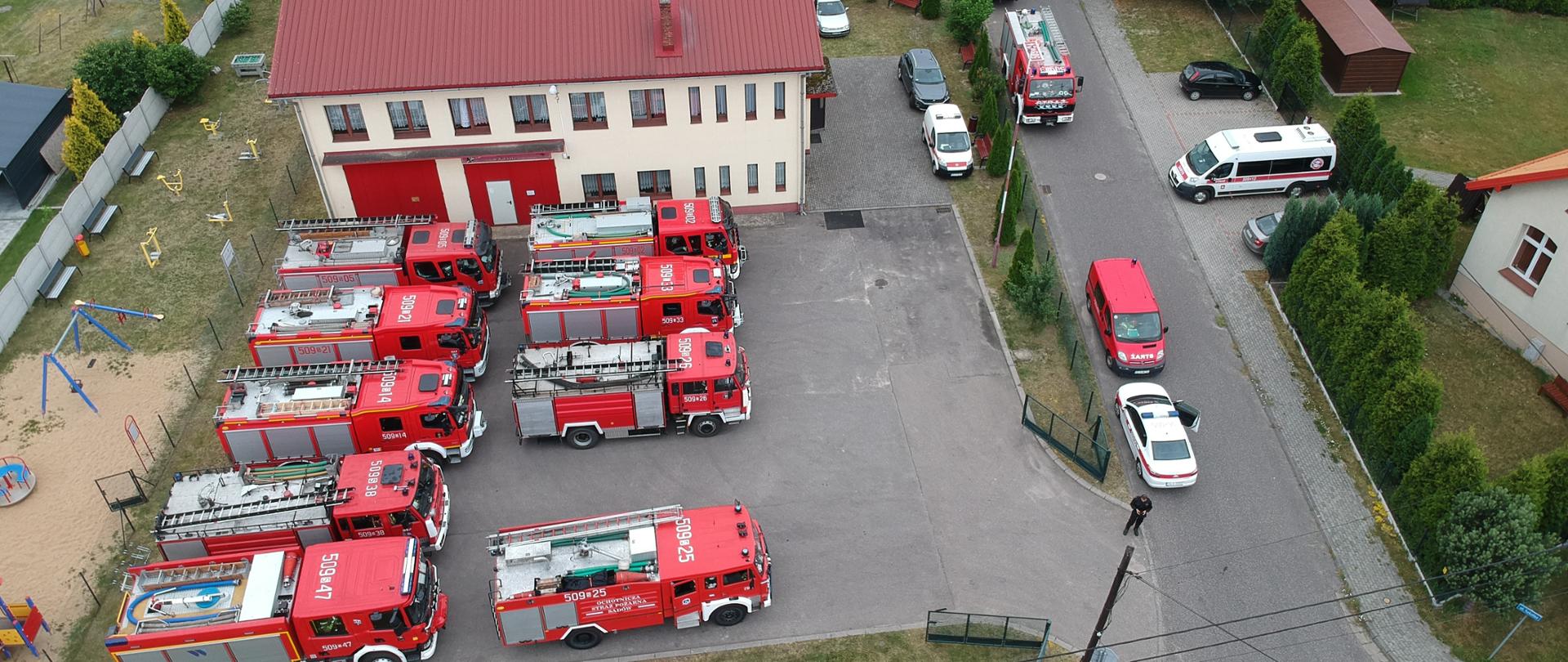 Zdjęcie zrobione z lotu ptaka przed budynkiem strażnicy. Przed budynkiem stoją zaparkowane wozy strażackie.