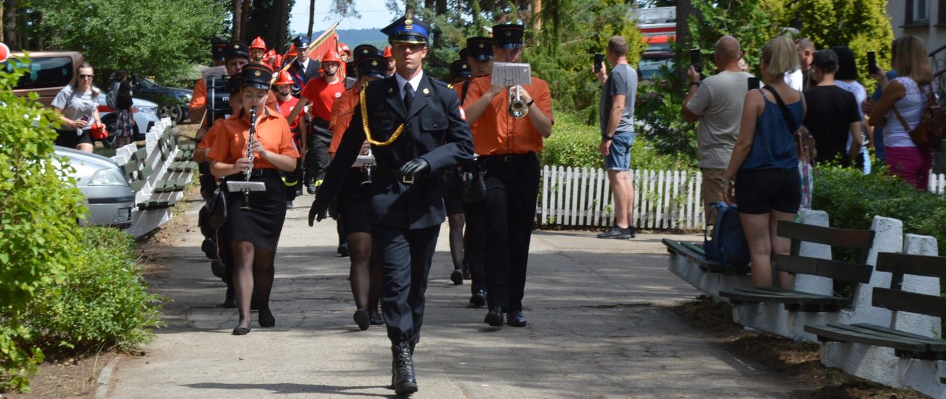 Zdjęcie przedstawia strażaka, który prowadzi pododdział marszowy. Za strażakiem jako pierwsza idzie orkiestra. Z boku stoją ludzie, którzy robią zdjęcia.