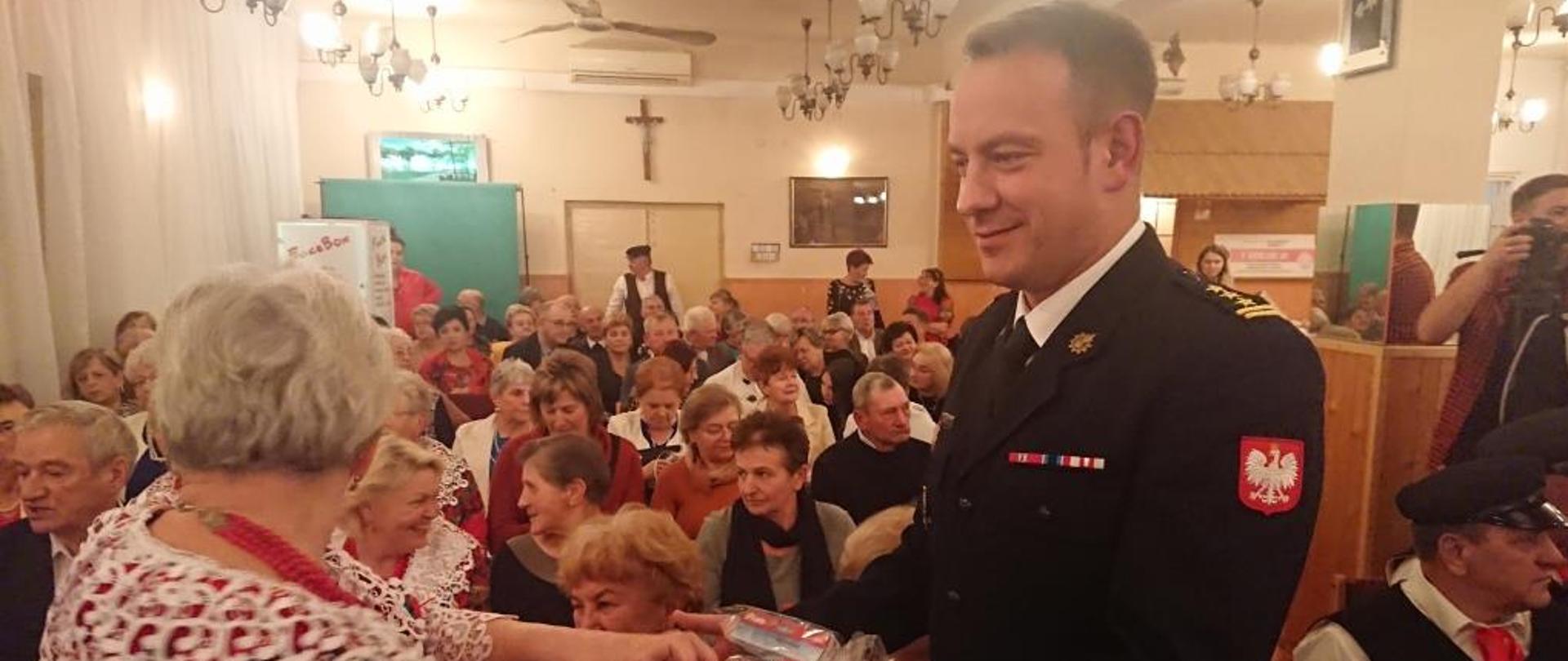 Zdjęcie wykonane na sali szkoleniowej Ochotniczej Straży Pożarnej w Jedlińsku. Na zdjęciu widać mieszkańców gminy Jedlińsk, przedstawicieli samorządu lokalnego wraz z wójtem gminy oraz członków jednostek ochotniczych straży pożarnych. Zgromadzeni uczestnicy biorą udział w pogadance tematycznej „czujka na straży twojego bezpieczeństwa” zorganizowanej w ramach obchodów VIII dnia seniora. Zgromadzone na sali osoby siedzą na krzesłach ustawionych w rzędach. 