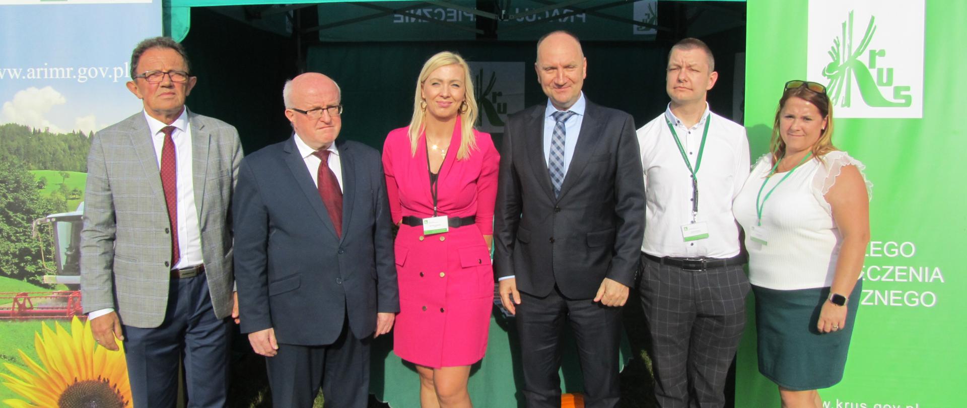 Czterech mężczyzn i dwie kobiety na tle zielonego namiotu z logiem KRUS i napisem Pracuj Bezpiecznie 