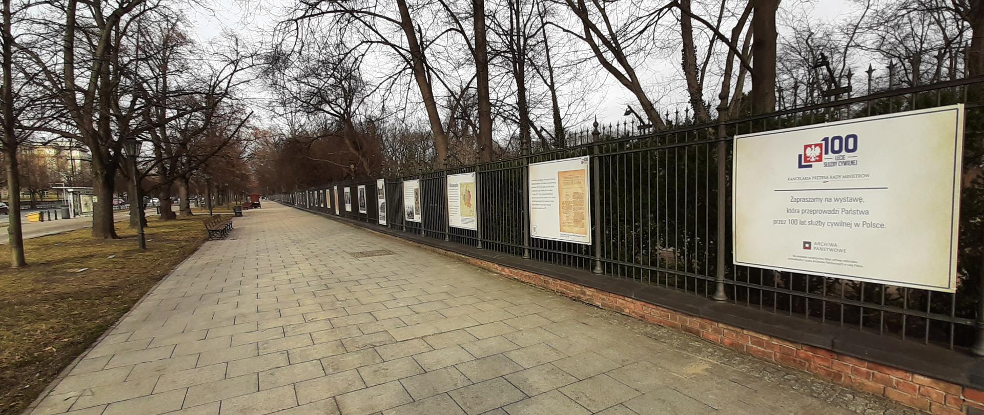 Na zdjęciu wystawa plenerowa z okazji 100-lecia służby cywilnej. Na zdjęjciu znajduje się chodnik wzdłuż Alej Ujazdowskich. Po lewej stronie od chodnika znajduje się pas trawy. Po prawej stronie chodnika ogrodzenie Łazienek Królewskich, a na tym płocie plansze z wystawą.