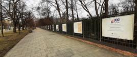 Na zdjęciu wystawa plenerowa z okazji 100-lecia służby cywilnej. Na zdjęjciu znajduje się chodnik wzdłuż Alej Ujazdowskich. Po lewej stronie od chodnika znajduje się pas trawy. Po prawej stronie chodnika ogrodzenie Łazienek Królewskich, a na tym płocie plansze z wystawą.