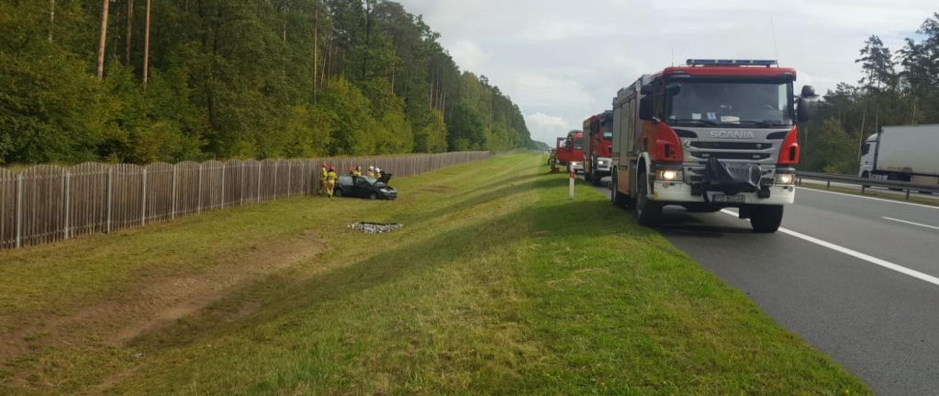 Dzień. Po prawej stronie widoczne trzy wozy strażackie stojące na pasie awaryjnym autostrady. Po lewej stronie uszkodzony samochód osobowy oraz grupa strażaków.