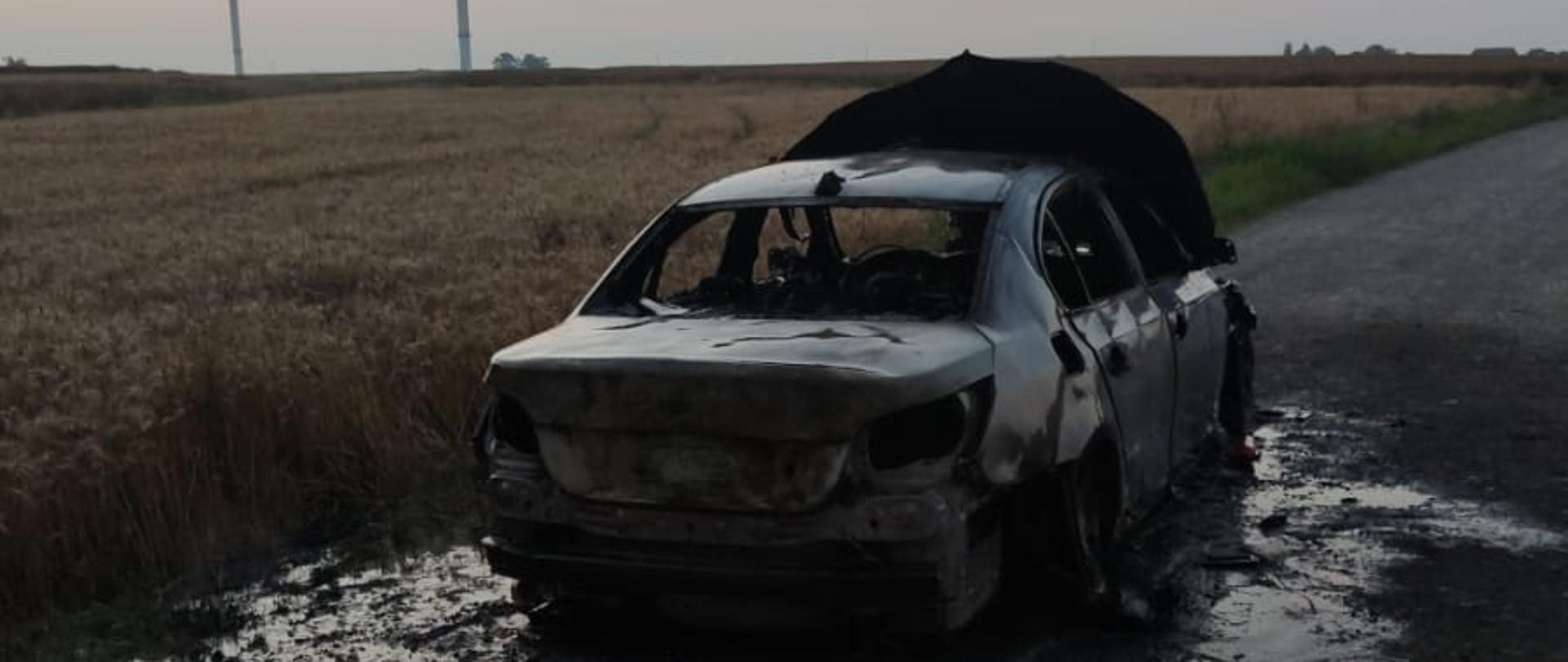 Na zdjęciu spalony samochód osobowy. W tle pole i wiatraki.