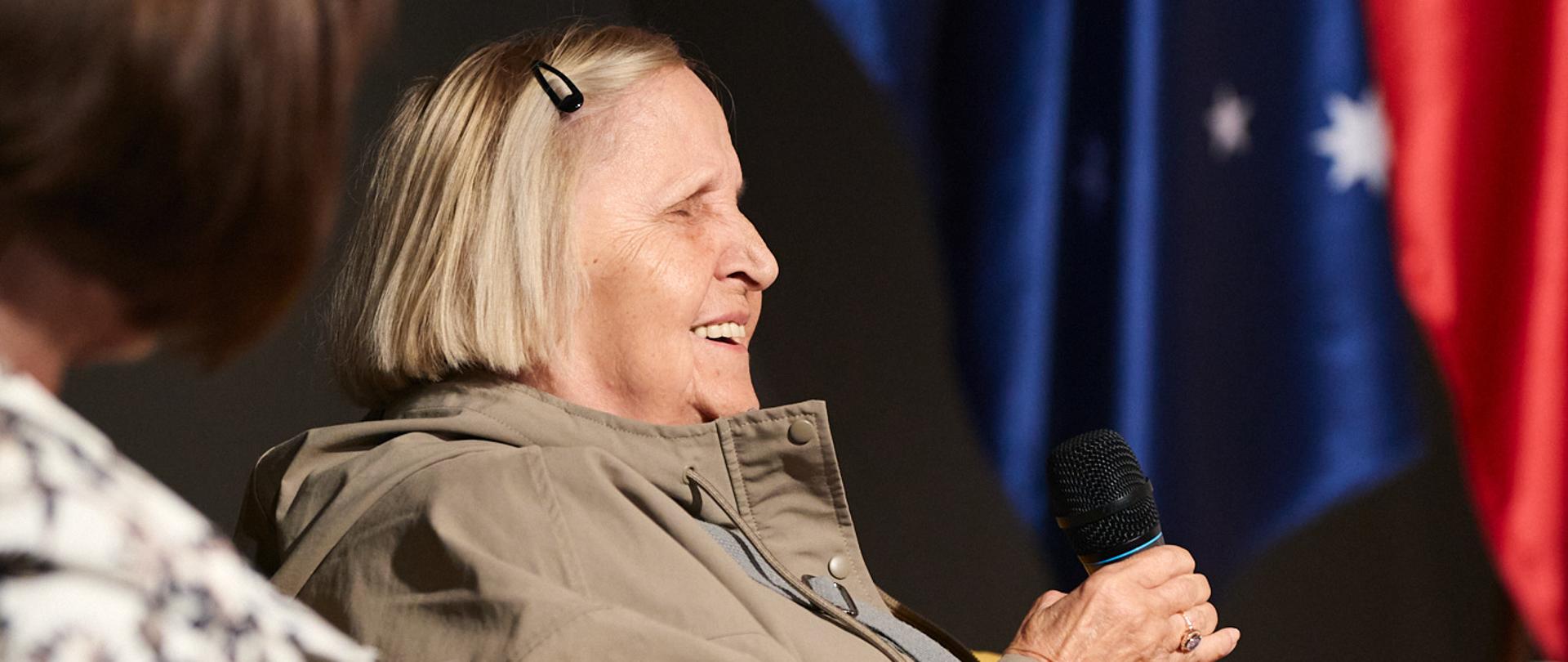 An elderly woman is holding a microphone and answering questions. In the background are the Australian and Polish flags.