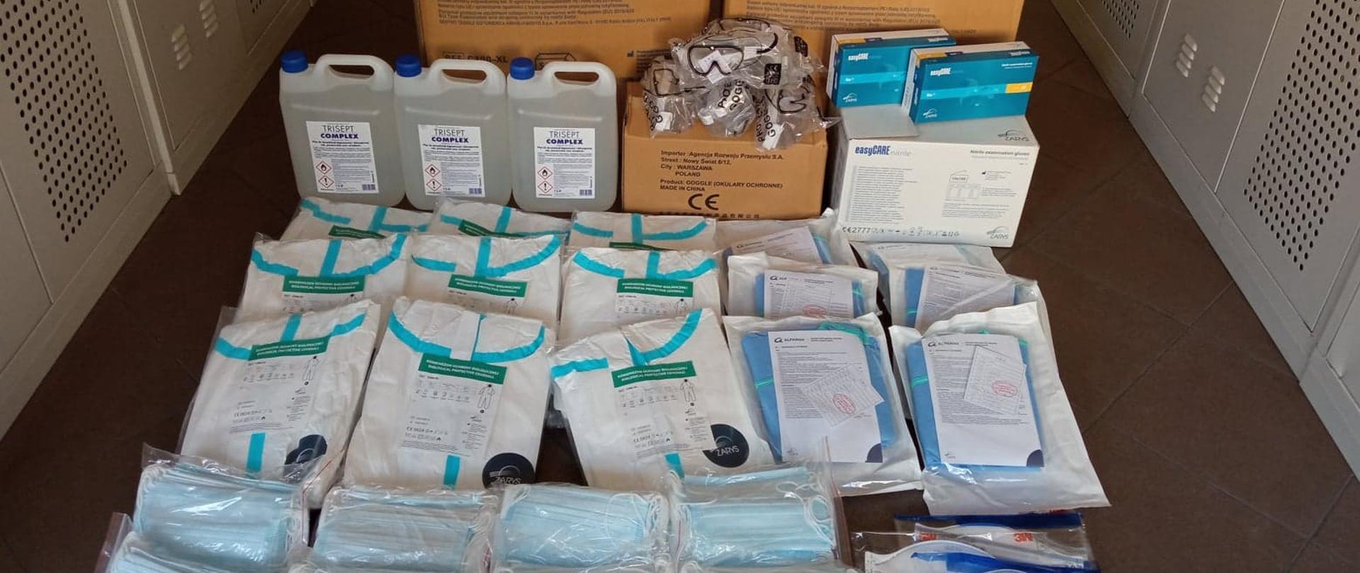 Środki ochrony indywidualnej dla ostrowskich strażaków. Na zdjęciu widoczne pojemniki ze środkiem dezynfekcyjnym, opakowania maseczek jednorazowych, kombinezonów ochronnych oraz rękawiczek jednorazowych.