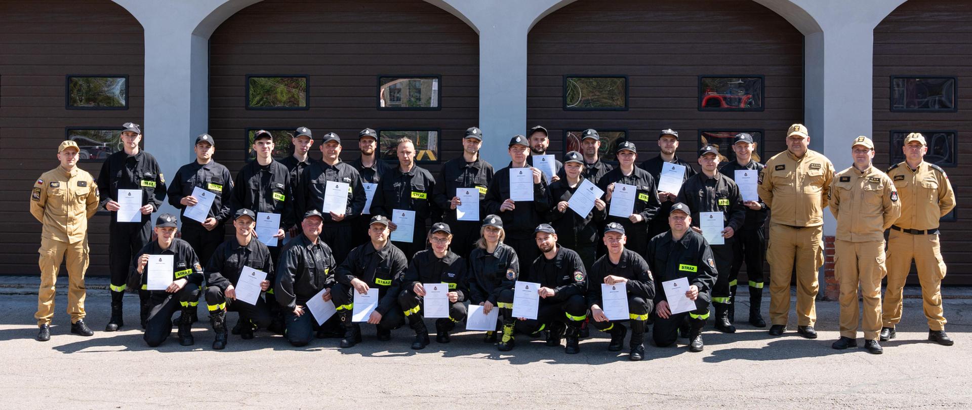 Wspólne zdjęcie komisji egzaminacyjnej wraz z druhami OSP po zakończonym kursie. Ochotnicy trzymają w rękach zaświadczenia. W tle bramy garażowe.