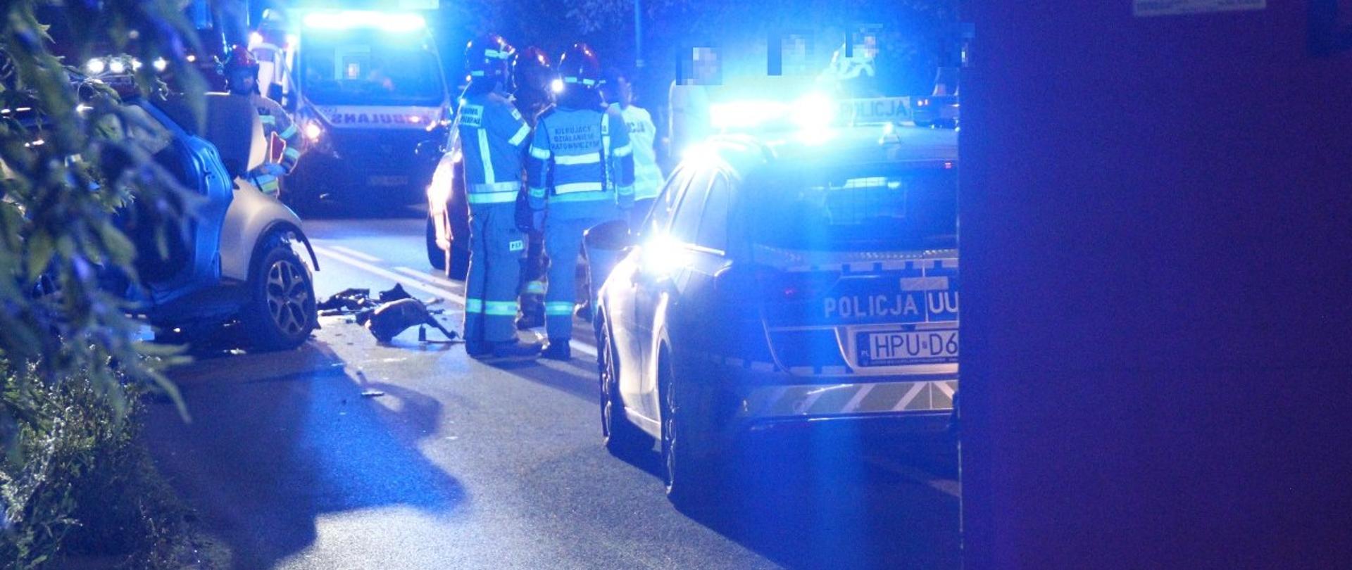 Zdjęcie przedstawia pojazdy służb ratowniczych stojących na drodze podczas obsługi interwencji, pojazdy mają włączone światła błyskowe koloru niebieskiego, na drodze stoją strażacy, którzy brali udział w likwidacji zagrożenia.