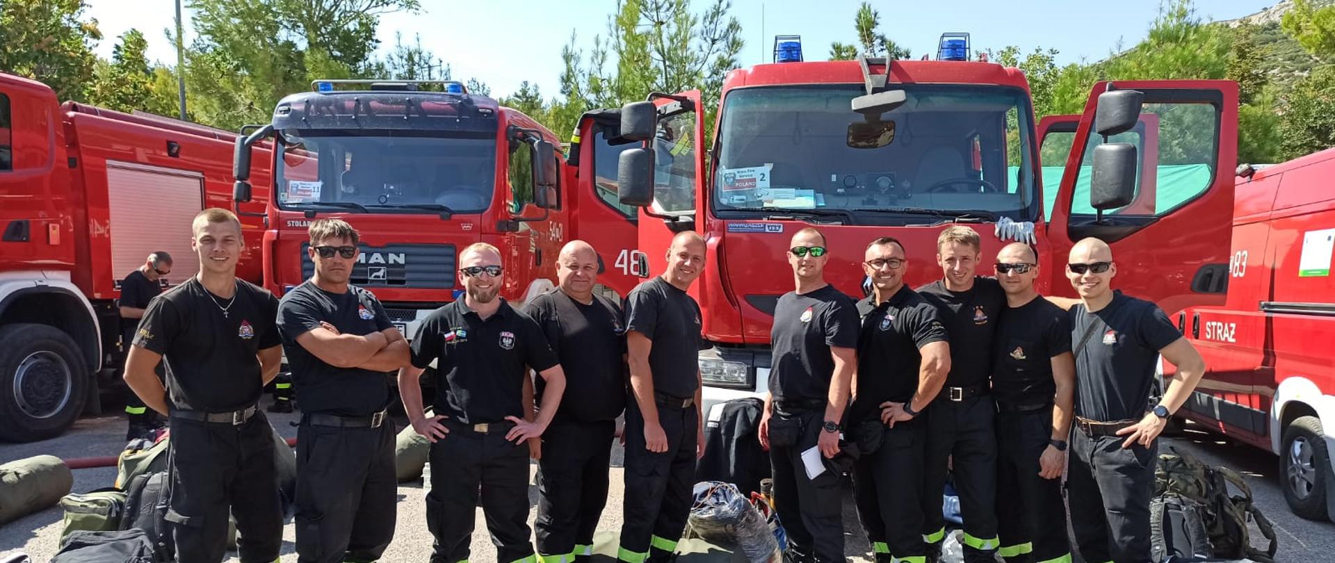 WYMIANA POLSKICH STRAŻAKÓW W GRECJI. Na zdjęciu widać kościańskich strażaków, którzy działali i tych którzy będą działali w Grecji, wokół nich torby podróżne, za nimi stoją samochody pożarnicze, w tle widać dzewa 