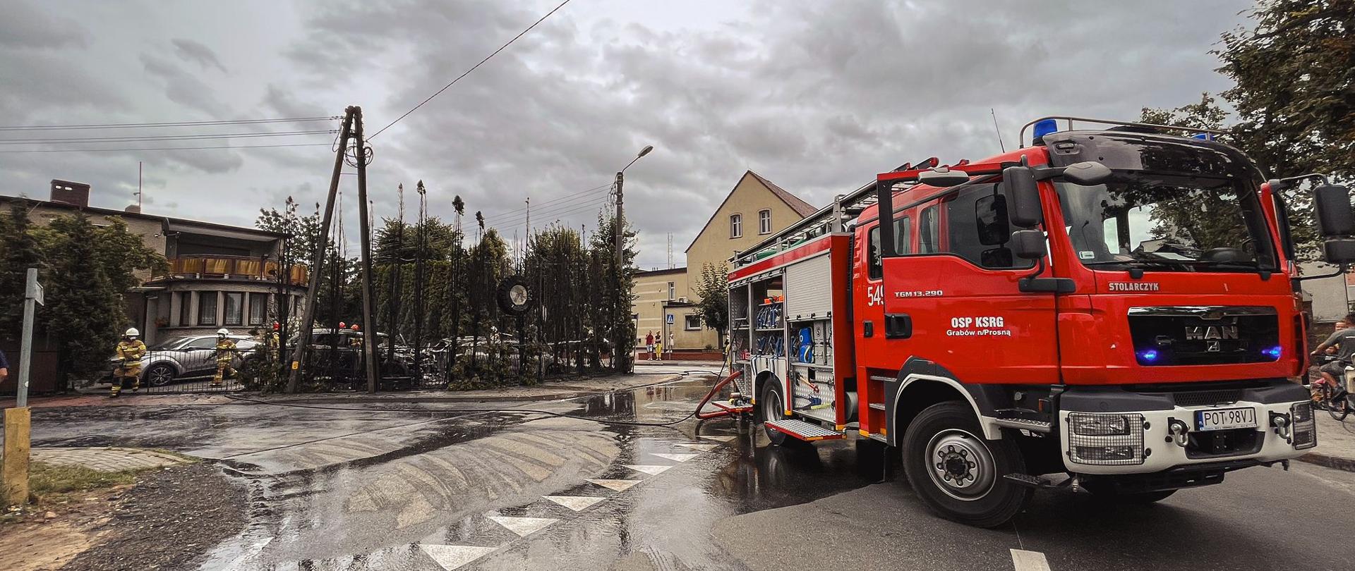 Samochód strażacki stoi na jezdni, na drugim planie spalone krzewy i samochody znajdujące się na terenie posesji, na której doszło do pożaru.
