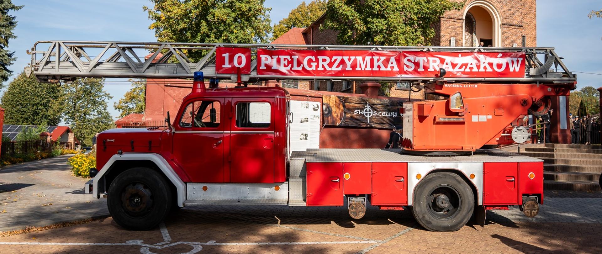 Czerwony, zabytkowy samochód pożarniczy z drabiną i banerem „10 PILGRZYMKA STRAŻAKÓW” zaparkowany przed Sanktuarium Maryjnym w Skoszewach.