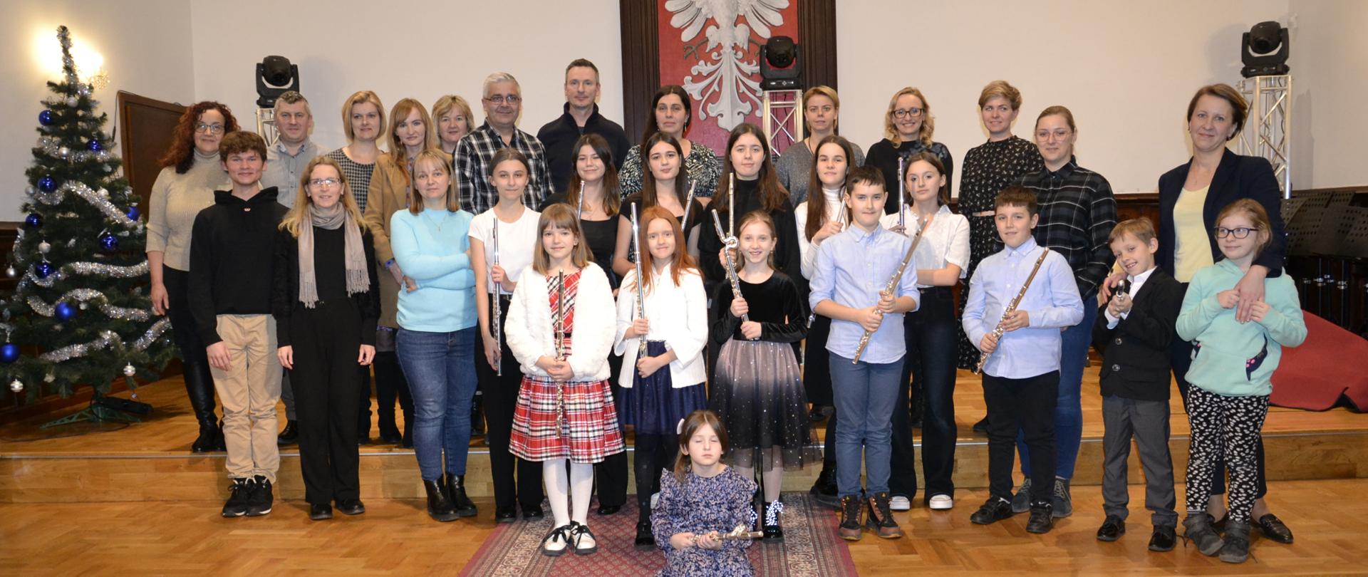 zdjęcie przedstawia grupę uczniów z klasy fletu PSM I i II stopnia w Mielcu oraz SM I stopnia w Niwiskach, którzy stoją na stopniu sceny na Sali Królewskiej PSM Mielec, po lewej stronie widać choinkę, w tle godło Polski oraz głowy oświetleniowe
